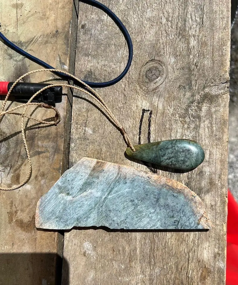 Pounamu roimata (teardrop) pendant on a braided cord beside a raw slab of stone, by Taonga by Timoti