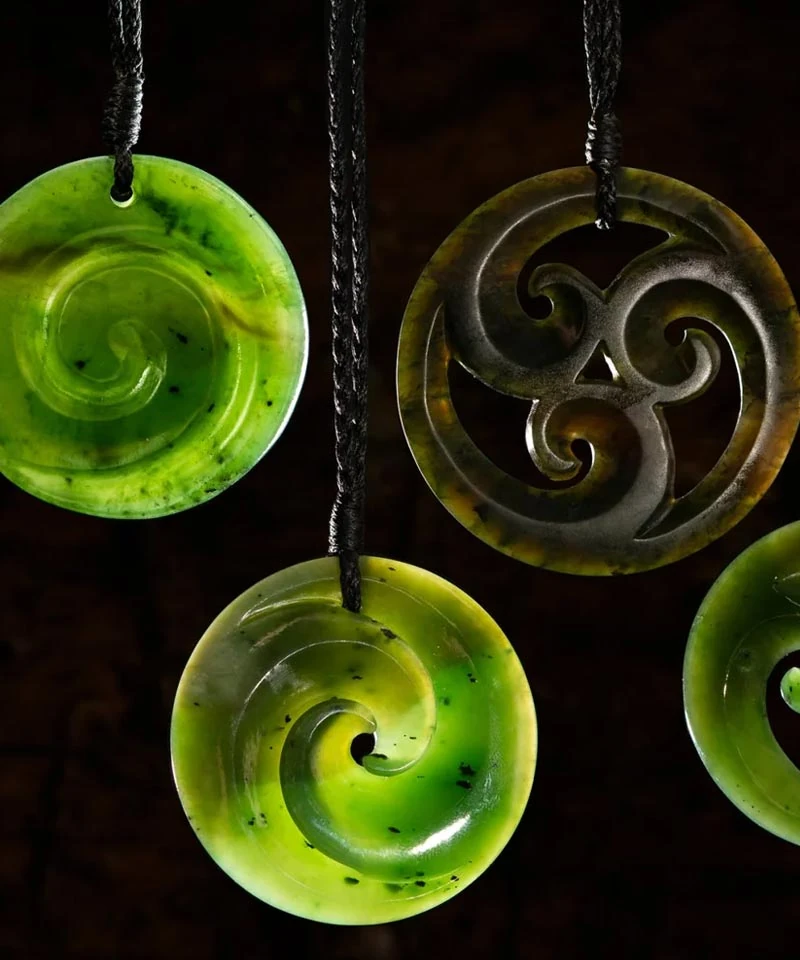 Round pounamu disc pendants carved with koru spirals, including one in darker stone with a triple koru design