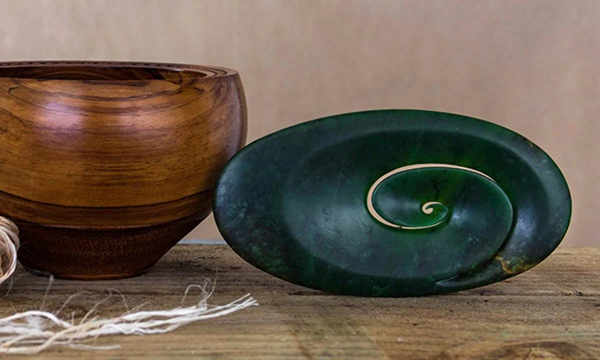 Carved oval pounamu piece with an open koru spiral beside a turned wooden bowl, at Hokitika Craft Gallery
