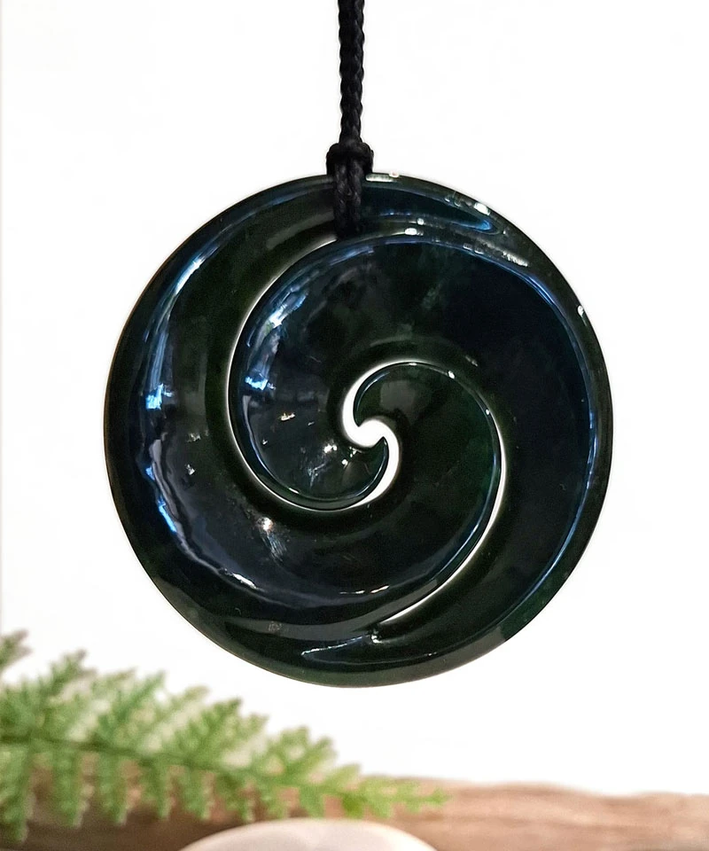 Dark polished pounamu disc pendant carved with a koru spiral on a black cord, from Hokitika Craft Gallery