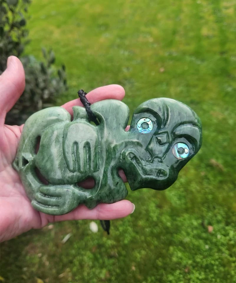 Pounamu hei tiki with pāua shell eyes held in an open hand, carved by Brett Phillips of Jade Art Carvings