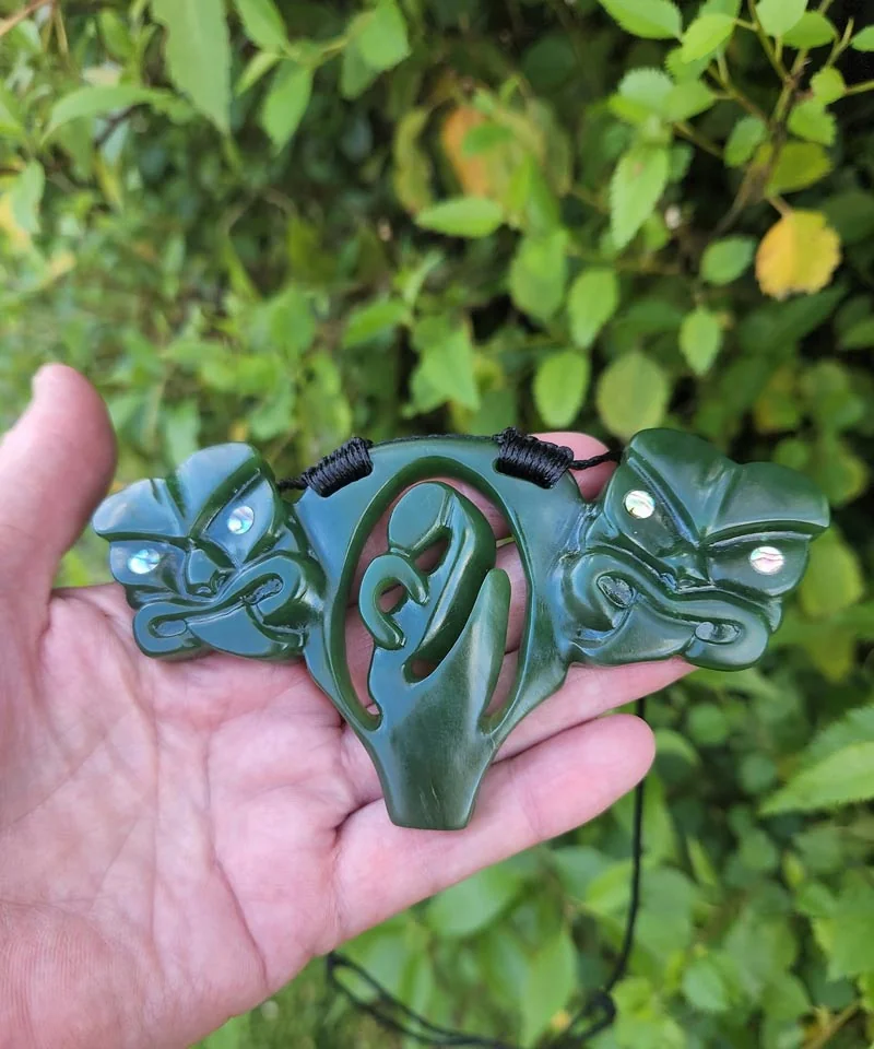 Pounamu pendant with two carved faces with pāua shell eyes framing a central koru, by Jade Art Carvings