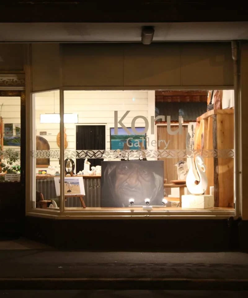 Koru Gallery shop window at night with a white carved stone hook sculpture, paintings and a portrait on display