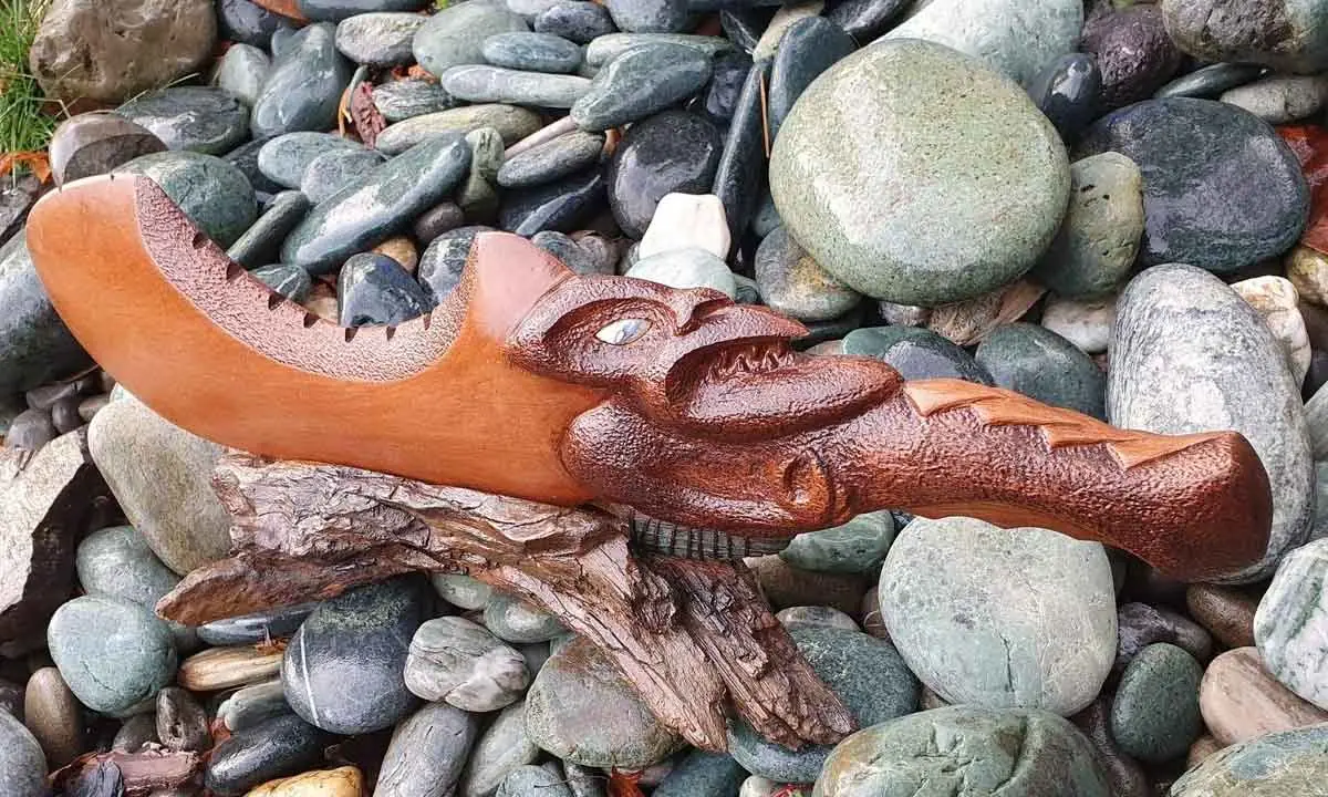Hand carved wooden club with a carved head and pāua shell eye resting on river stones, by Little Shop of Taonga