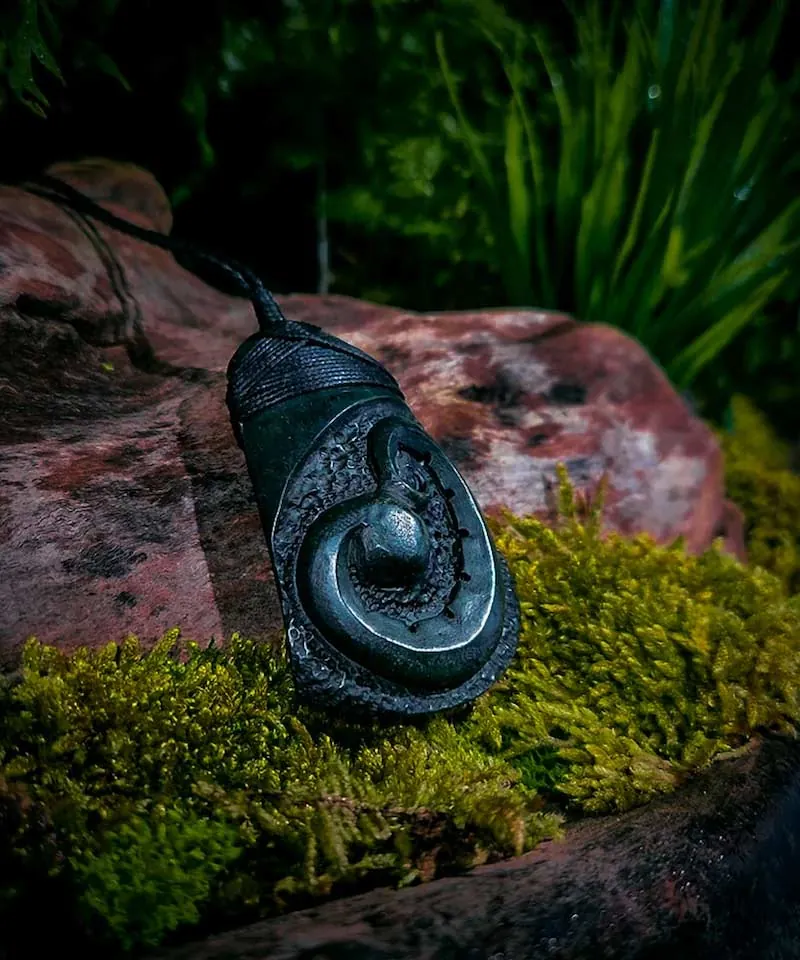 Dark stone pendant carved with a textured koru spiral and lashed with black cord, by Little Shop of Taonga