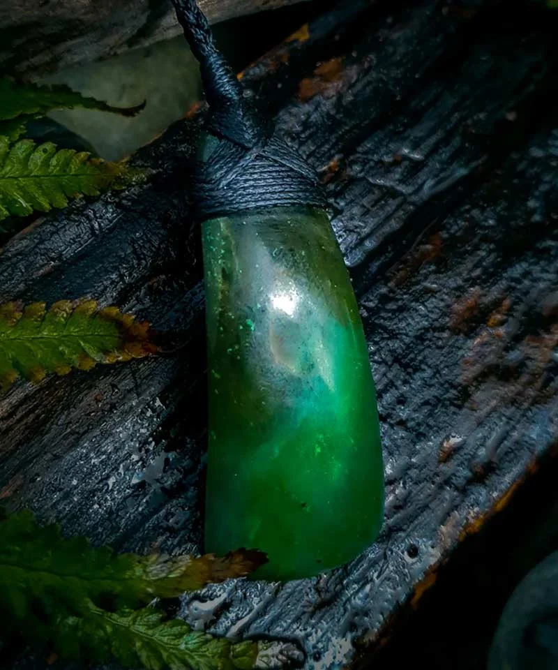 Bright green pounamu toki pendant lashed with black cord, resting on charred wood beside ferns, by Little Shop of Taonga
