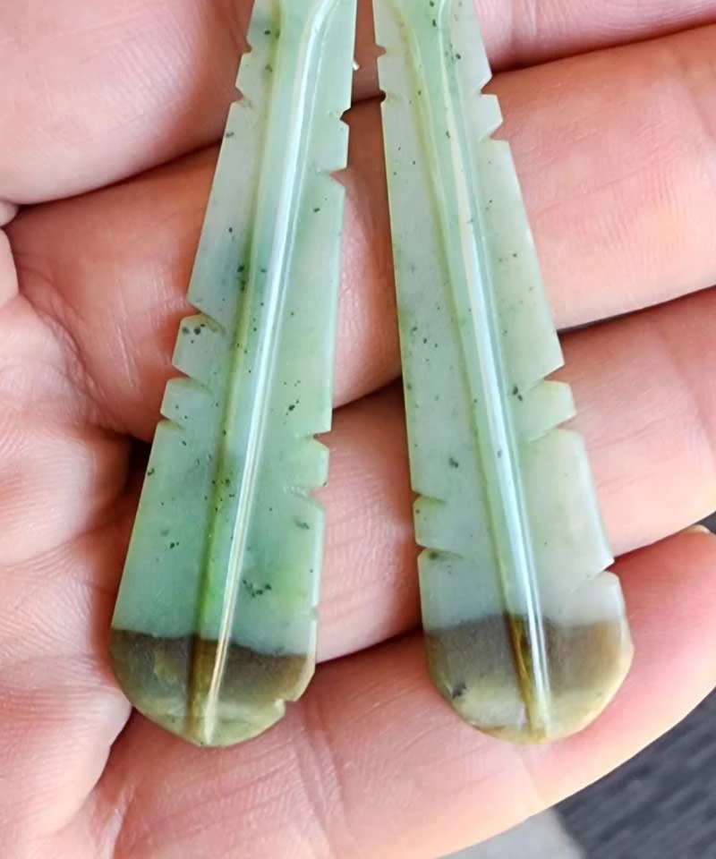 Pair of pale green jade feather earrings with brown tips held in a hand, carved by Thompson Carvers