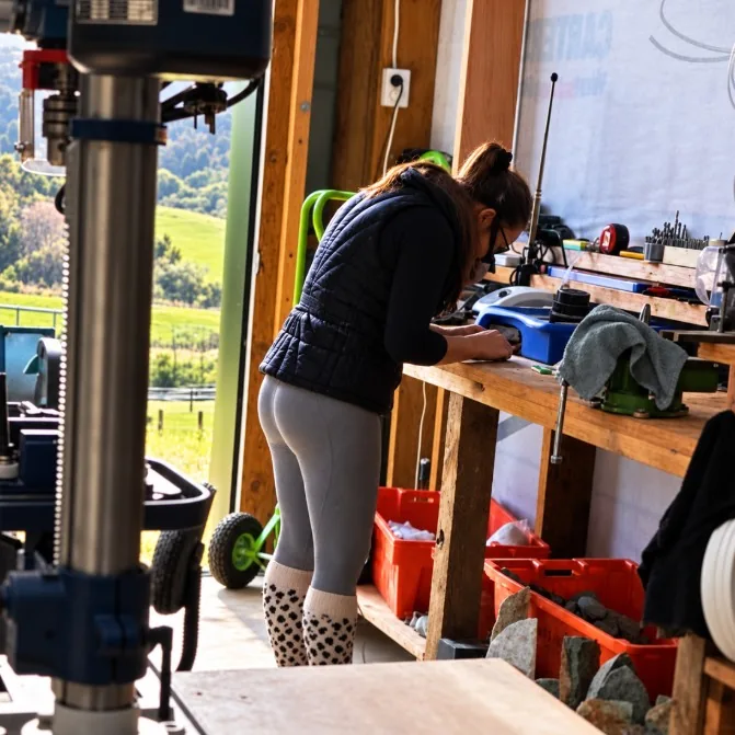Antoinette working at her carving bench in her Kerikeri workshop, with crates of raw stone and farmland outside