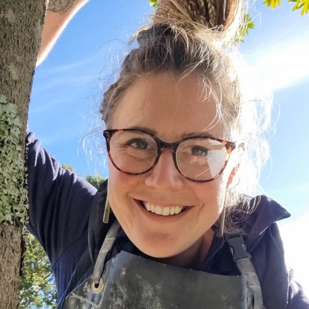 Antoinette smiling in her stone-dusted carving apron under a tree on a sunny day