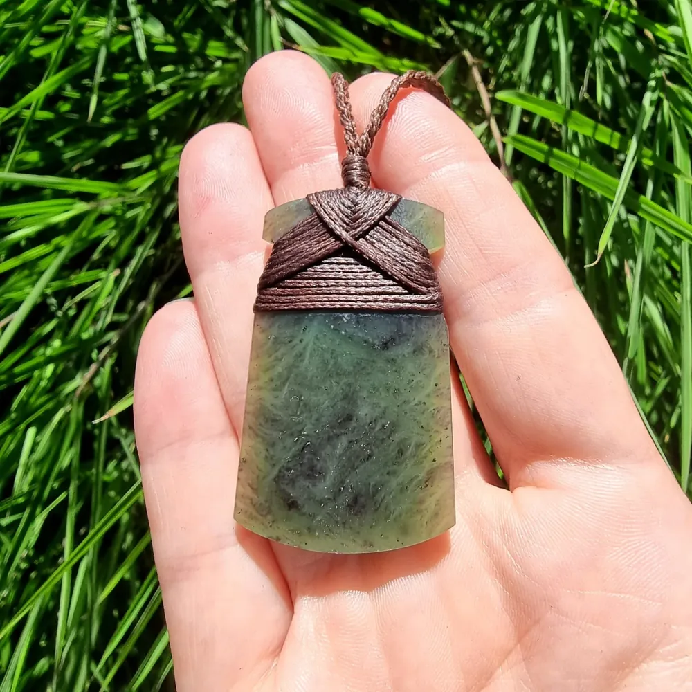 Pounamu hei toki pendant with brown bound cord held in an open hand over green grass